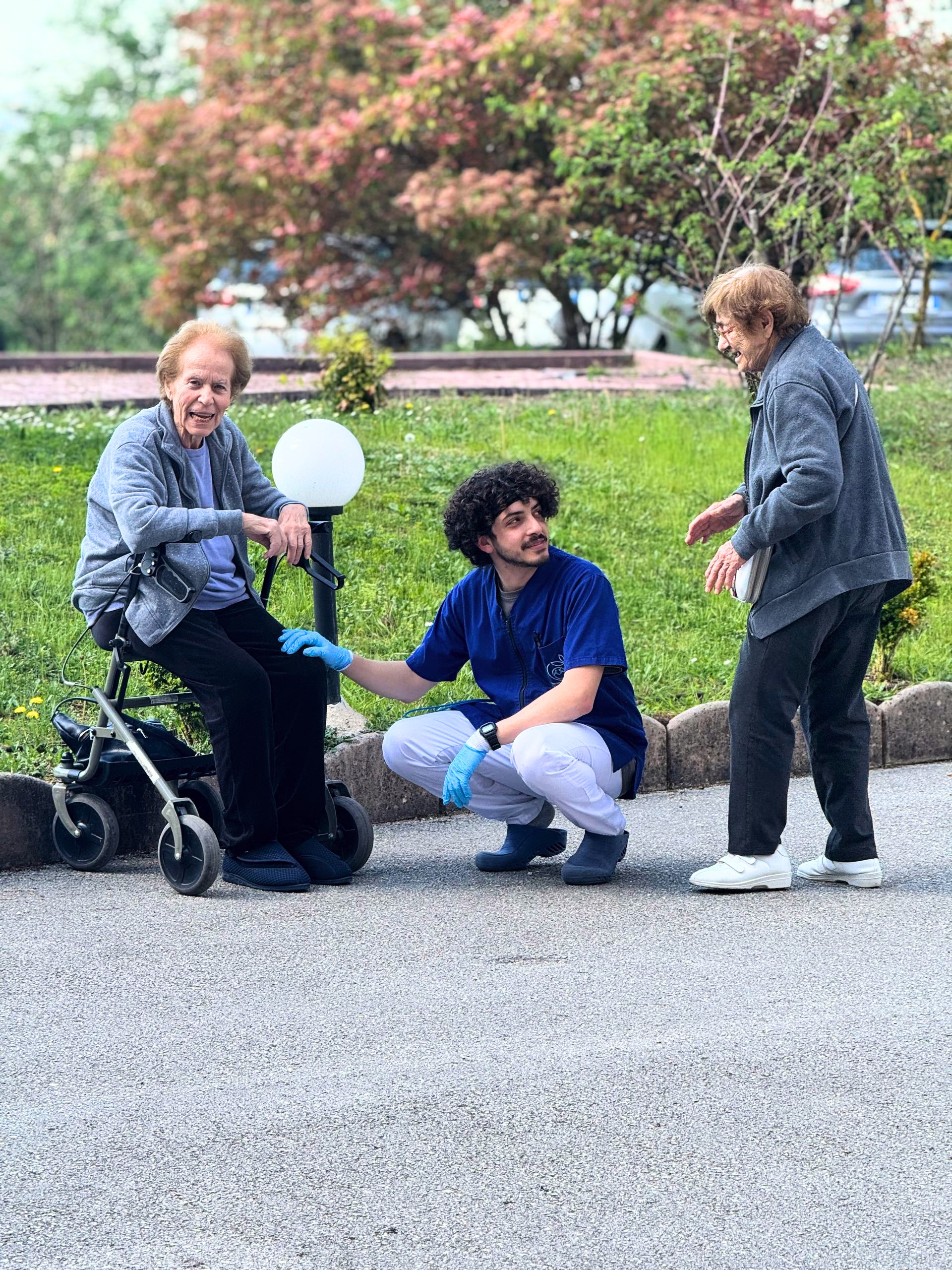 Un operatore di Villa Salmata accovacciato sorride accanto a due ospiti anziane nel giardino della struttura